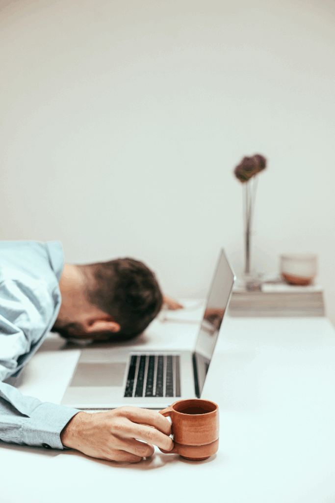 Exhausted man collapsed over laptop, holding a coffee mug - representing digital overwhelm.