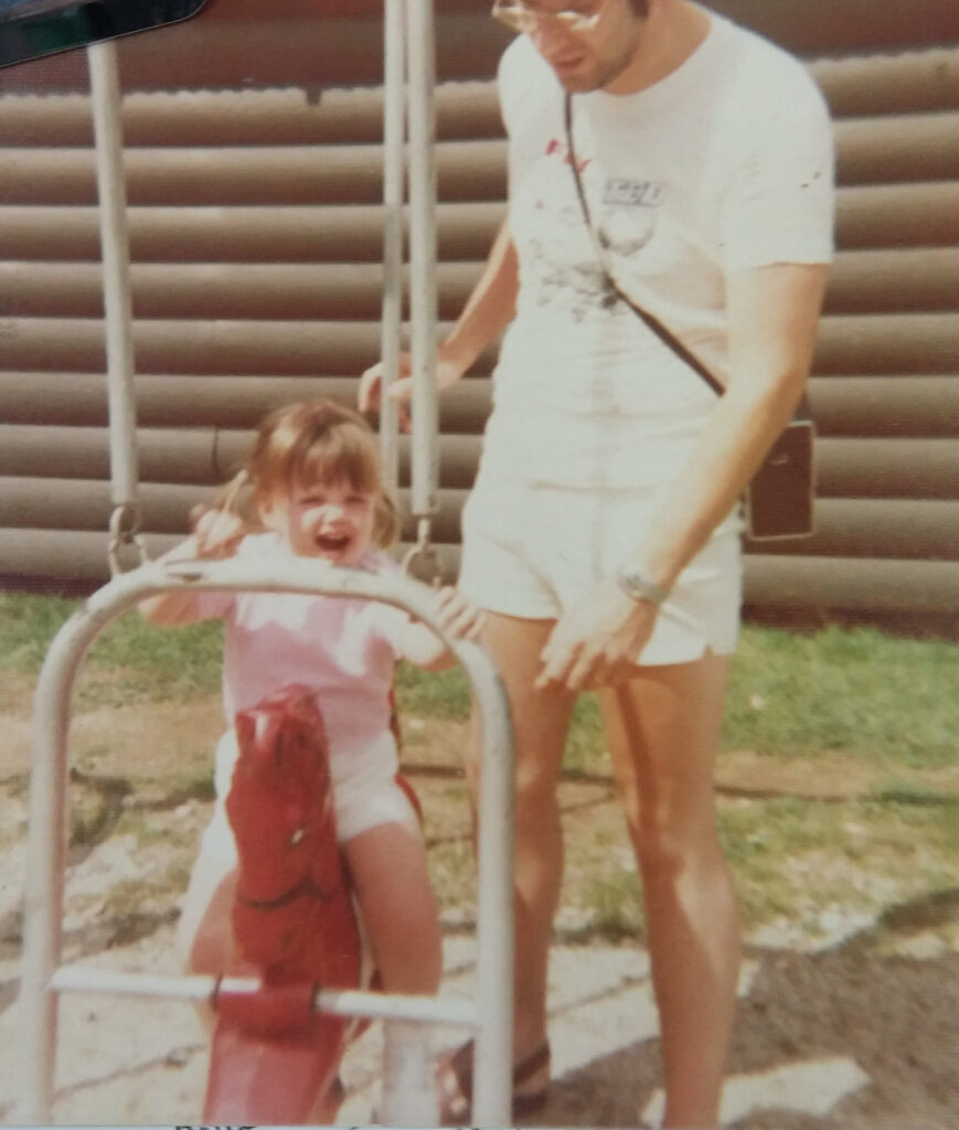 Jennifer as a child on a playground ride with father standing beside her.f