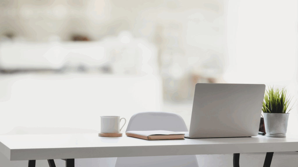 Minimalist white desk setup with laptop, notebook, coffee mug, and plant - symbolizing calm, organized digital work.